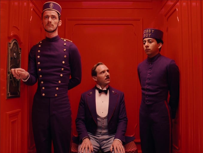 In the red lift of a hotel, a distinguished British gentleman and a swarthy lobby-boy have a conversation while the liftman looks on, all dressed in purple uniform