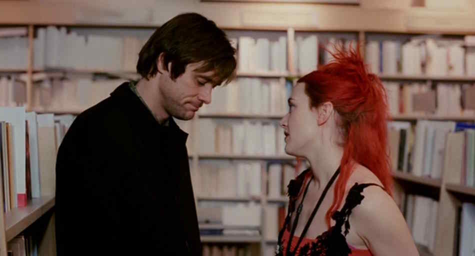 A young white man with black hair and a young white woman with dyed red hair look longingly at one another in a bookstore whose covers are fading away