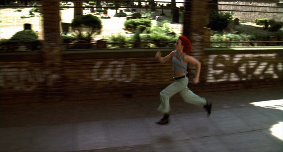 A young white woman with vibrant red hair, dressed in a ’90s-ass tracksuit, runs across the streets of Berlin