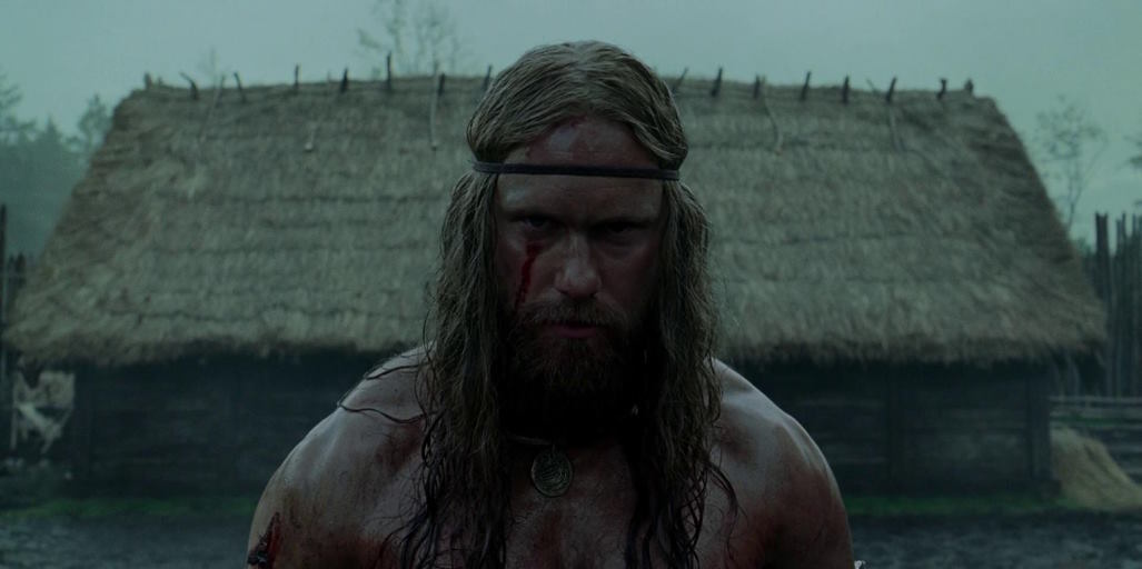 A viking, blood on his face, looks on sternly while raiding a village