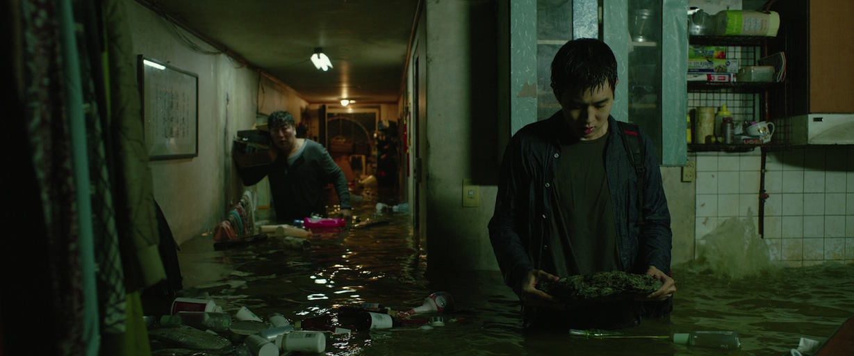 A young East Asian man looks at the rock he is holding while his father busily tries to rescue items behind him in their flooded house