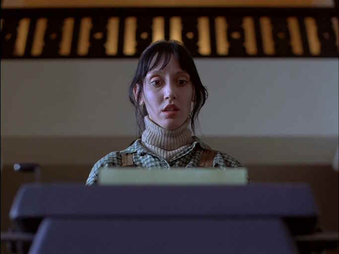 A concerned white woman of middle age with untidy strands of black hair, wearing a jumper, looks in horror at a typewriter whose contents we do not see