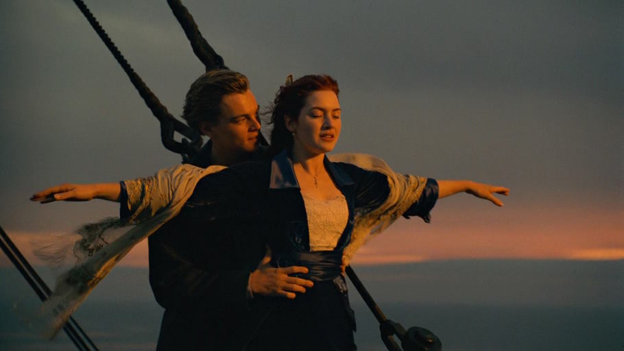 A young, handsome, clean-shaven white man with trim blonde hair holds a young white brunette in fancy clothes as she puts her arms out standing on the deck of a ship while the sun sets behind them